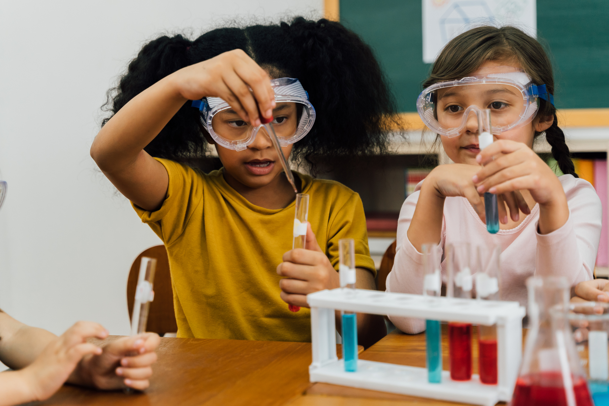 Multiethnic girls in safety goggles sitting at table mixing liquids in vials during chemistry lesson at primary school